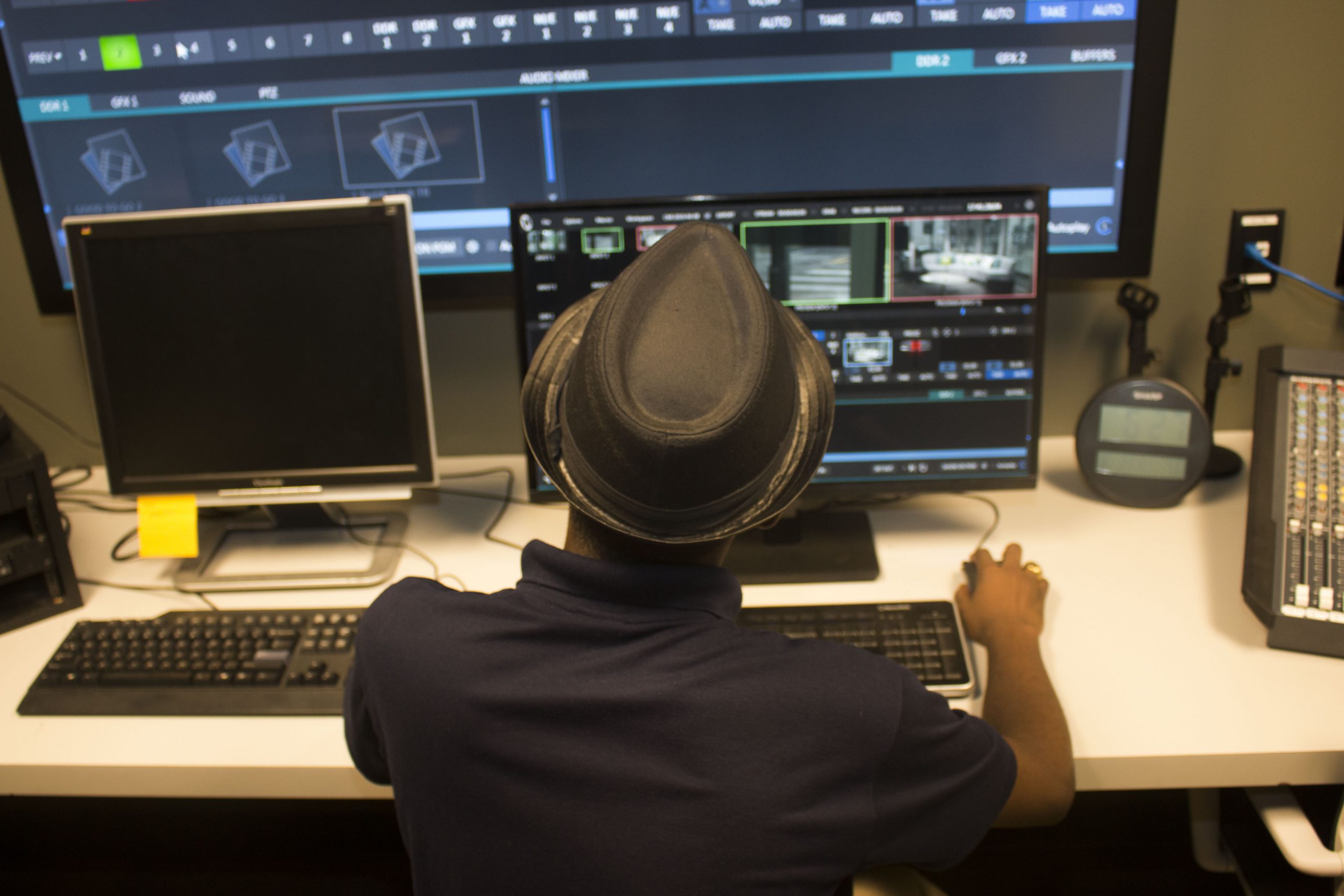 A person photographed from behind with a black hat and black shirt editing in the Center for Media Innovation's control room.