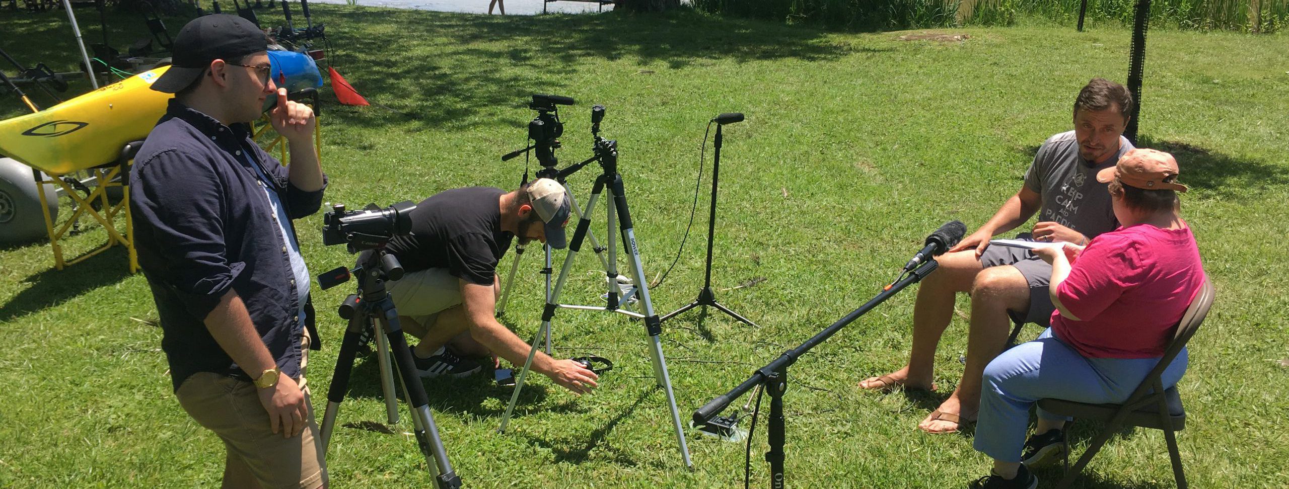 Photo of a group outside. two people are sitting in conversation with microphones pointed at them, one is setting up video equipment and one is standing. They are in a field by a lake.