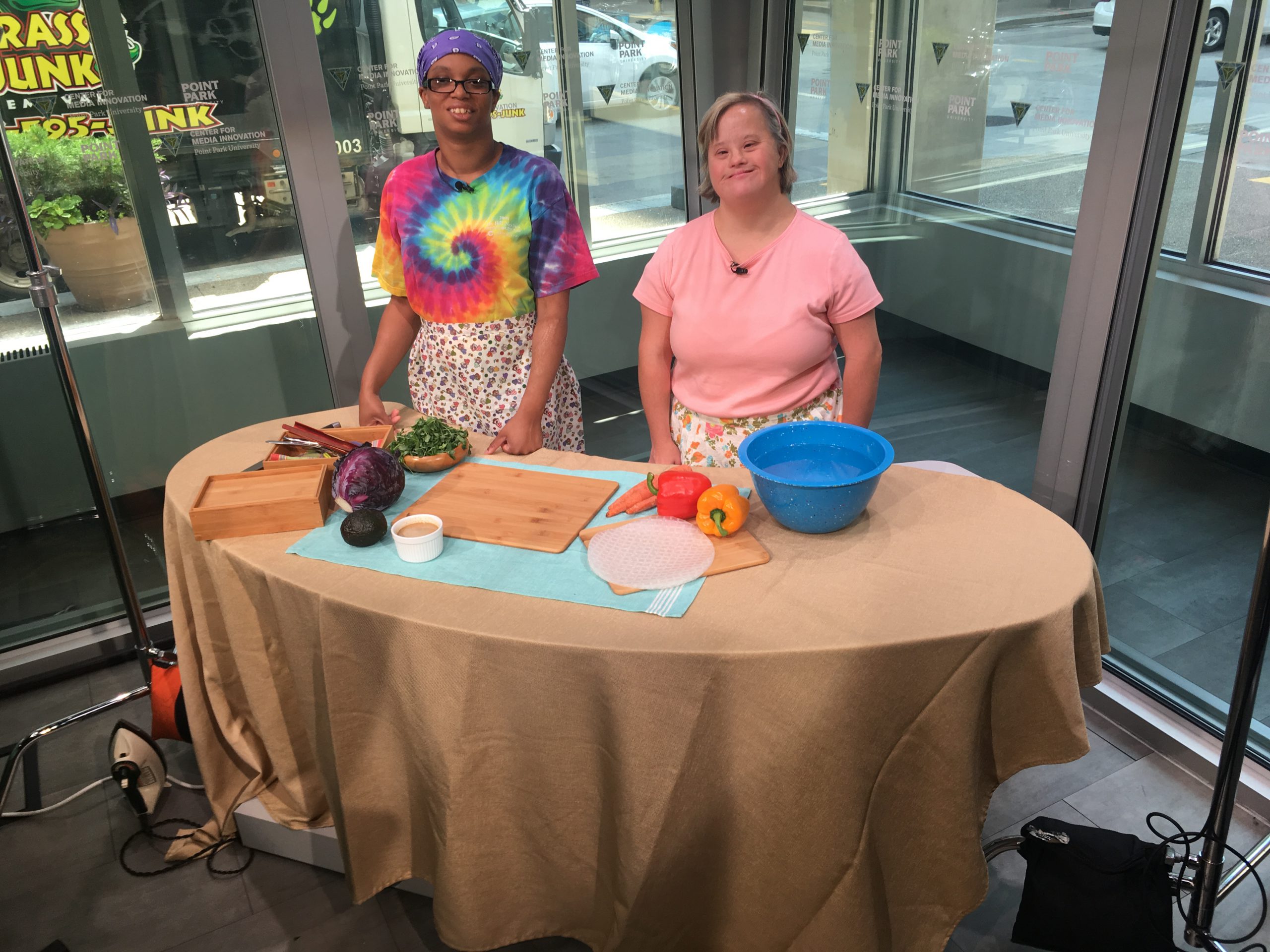 Two women stand behind a table with food ingredients on it, in the Center for Media Innovation's television studio. One woman is Black with black hair pulled back with a bandanna, and Is wearing a multi-colored tie-dye t-shirt and an apron. The other woman is white with short blonde hair, wearing a pink, short sleeved top, and an apron.